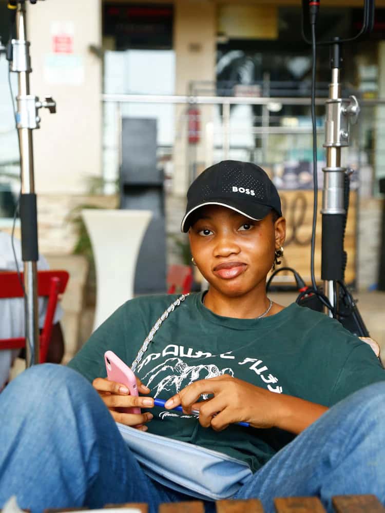 A Nigerian student in a cap holding a phone and a pen, reviewing lesson notes.